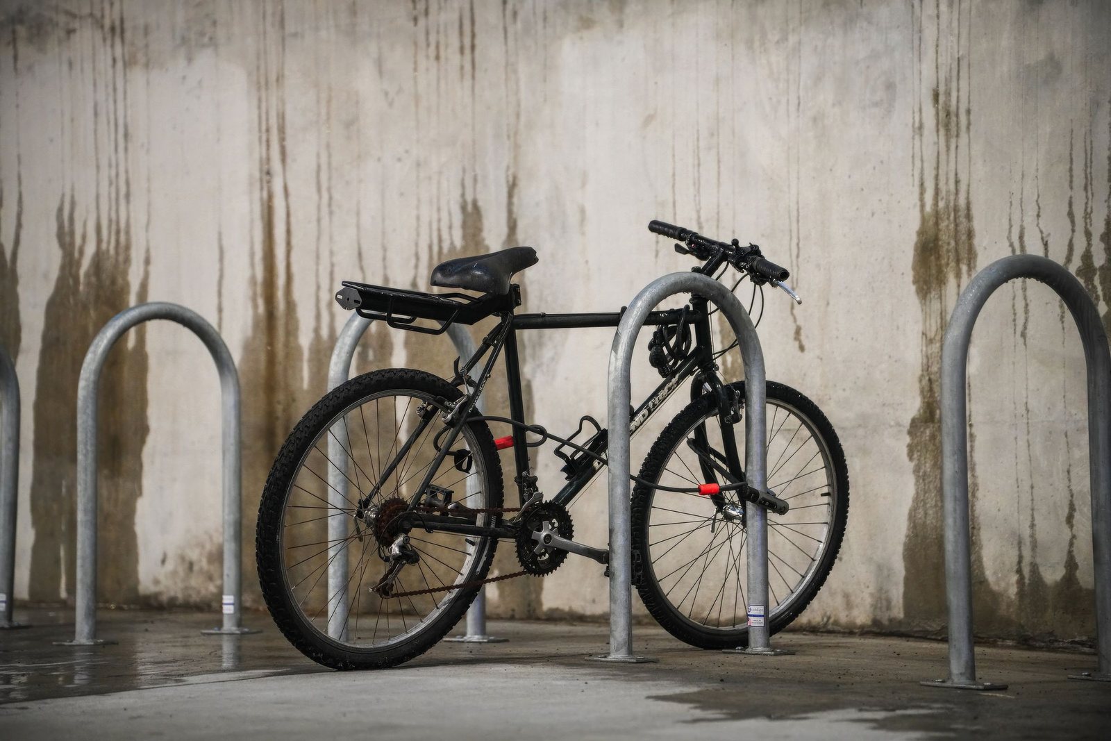 Black bicycle secured with a cable lock to a metal bike rack against a weathered concrete wall