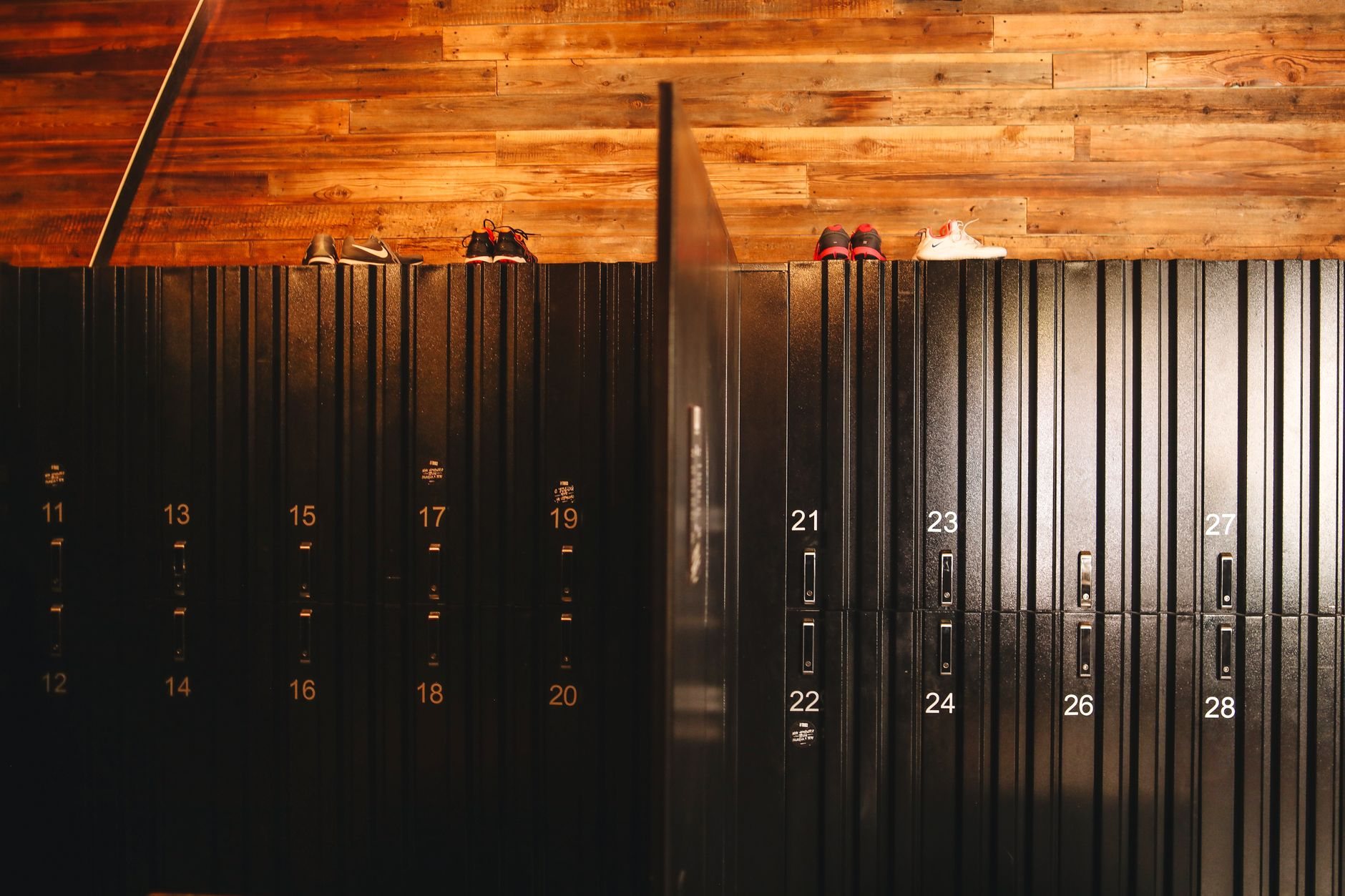 A bank of numbered black metal lockers in a gym changing room with a reclaimed wood wall and training shoes on top