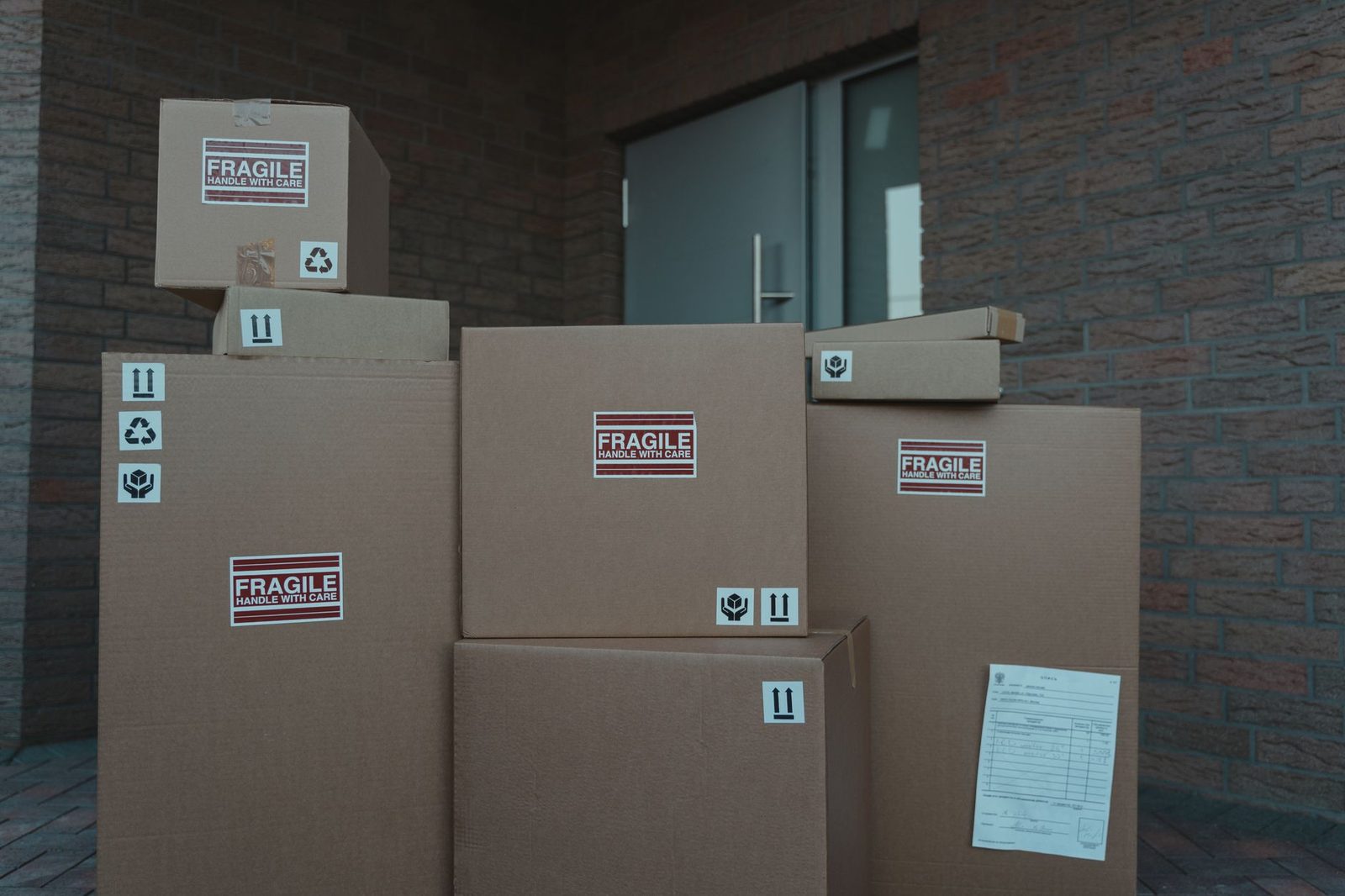 Stacked cardboard delivery parcels marked fragile piled outside a glass building entrance door in a brick vestibule