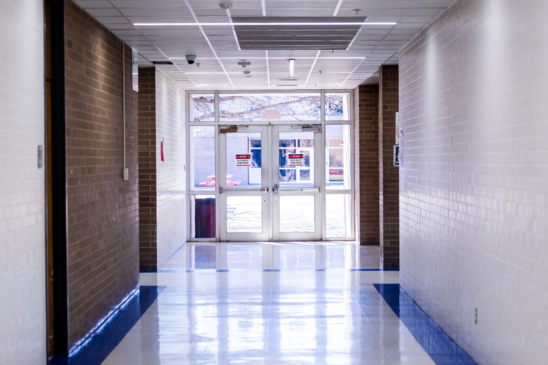 A school corridor leading to a pair of aluminum exit doors fitted with overhead door closers and do-not-enter signage