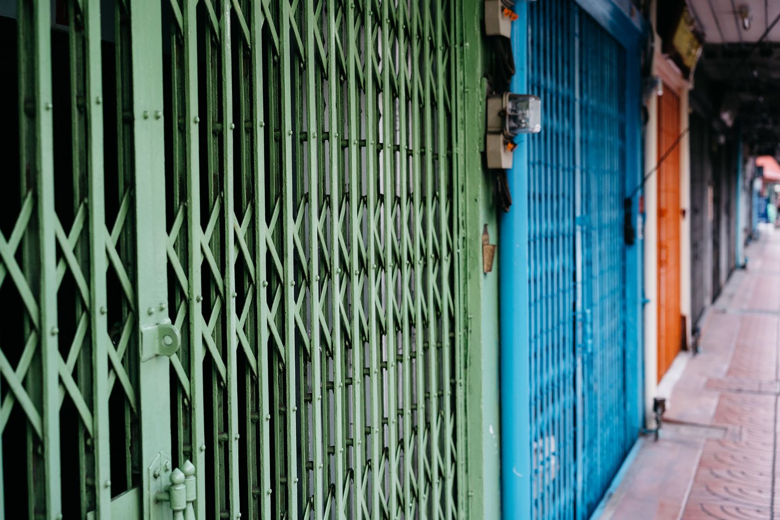 Row of closed accordion security gates on a street of shops, with the keyed lock plate visible on the leading edge of the nearest gate