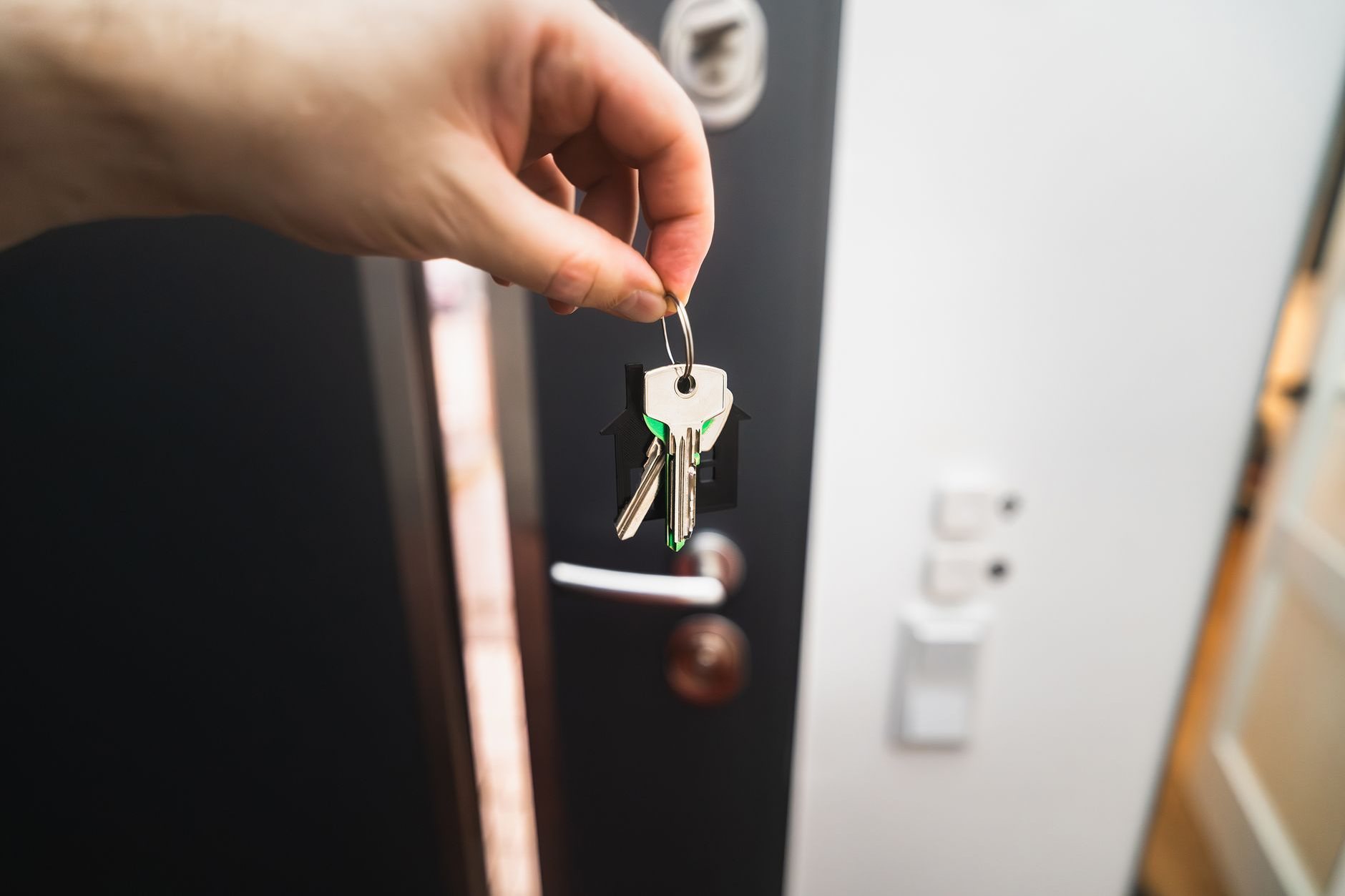 A hand holding a set of door keys in front of an open apartment door fitted with a deadbolt and a lever handle
