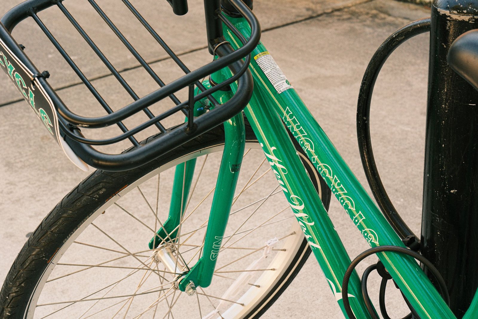 Close-up of a green bicycle locked with a cable to a black metal street post on a city sidewalk