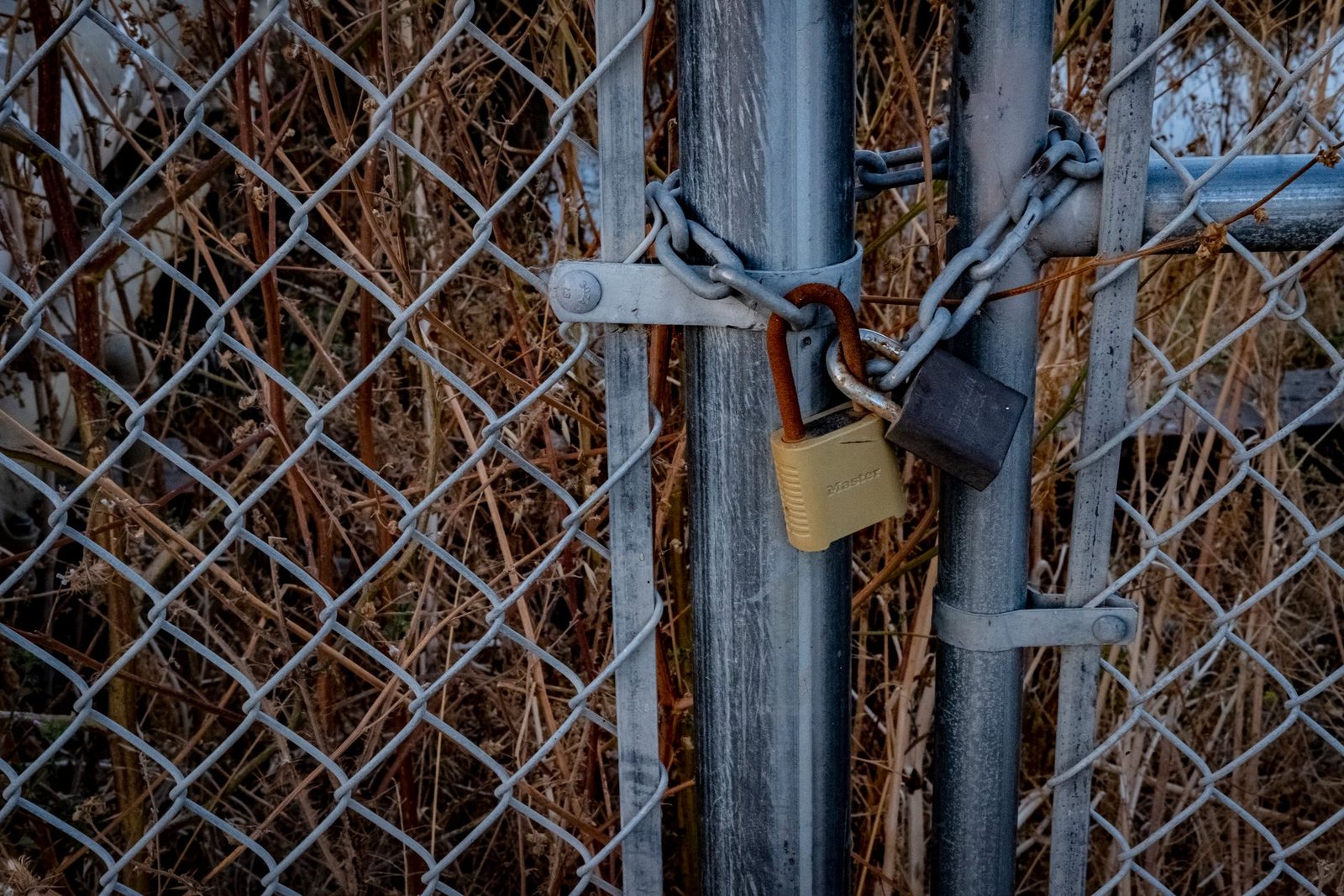 Two padlocks and a chain securing a chain-link gate, one shackle visibly rusted