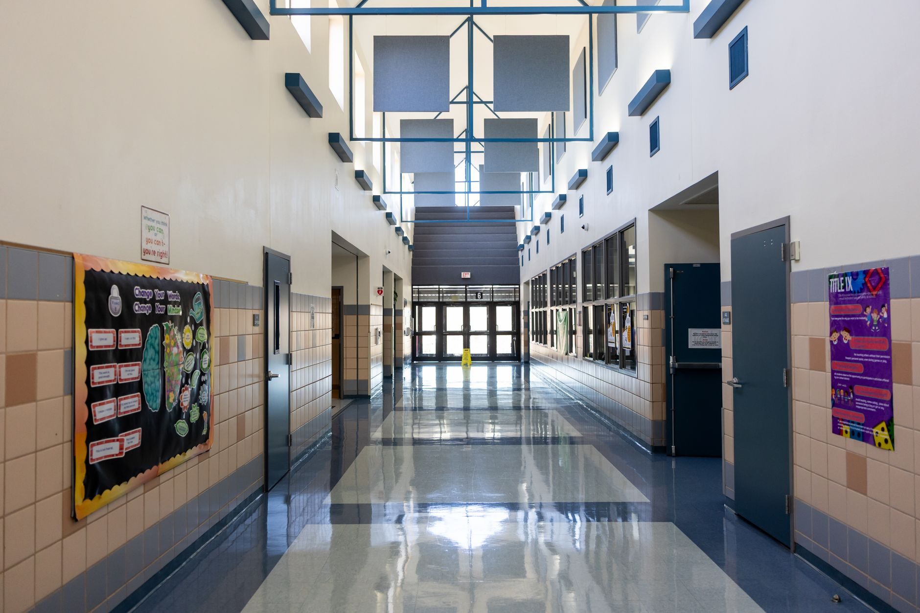 A bright school corridor lined with classroom doors fitted with lever handles and vision panels, leading to a glazed exit at the far end