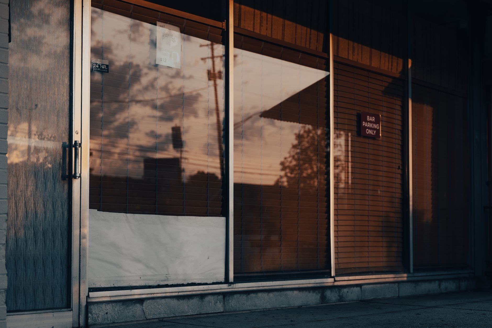 A closed storefront with aluminum-framed glass panels and an aluminum entry door at the left, photographed from the sidewalk at dusk